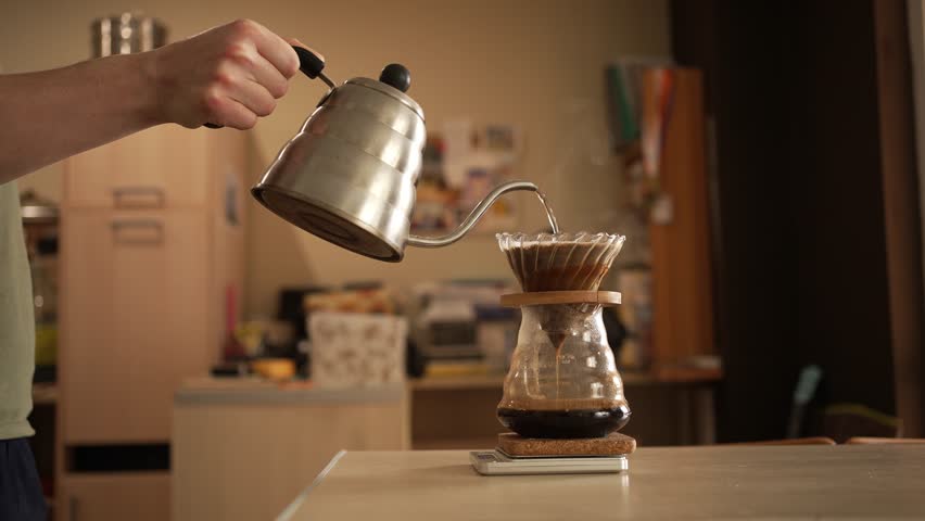 Professional barista carefully weighing and pouring hot water from copper kettle onto freshly ground coffee beans using manual pour-over brewing method in sunlit kitchen workspace