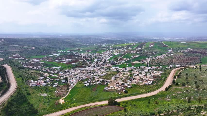 Aerial footage of a  hillside Arab village in Israel with dense cluster of flat-roof homes, winding roads, and natural mountainous terrain.  view showcases Middle Eastern Arab living in Israel .