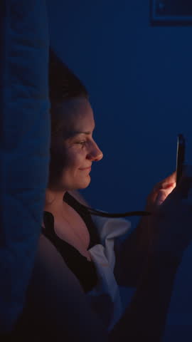 Vertical video. Young Woman Falls onto Pillow and Starts Typing a Message, Sends It and Hugs Phone to Chest with Anticipation, Captured from a Side Angle with Shallow Depth of Field, Romantic Communic