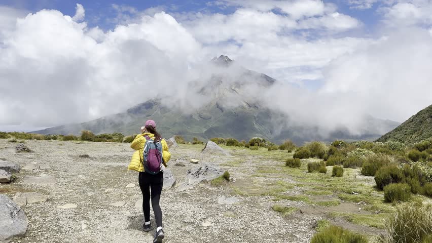 A girl in a yellow jacket hikes the Pouakai tarn trail past the mighty Mount Taranaki volcano in Egmont National Park. Spectacular trails on New Zealand