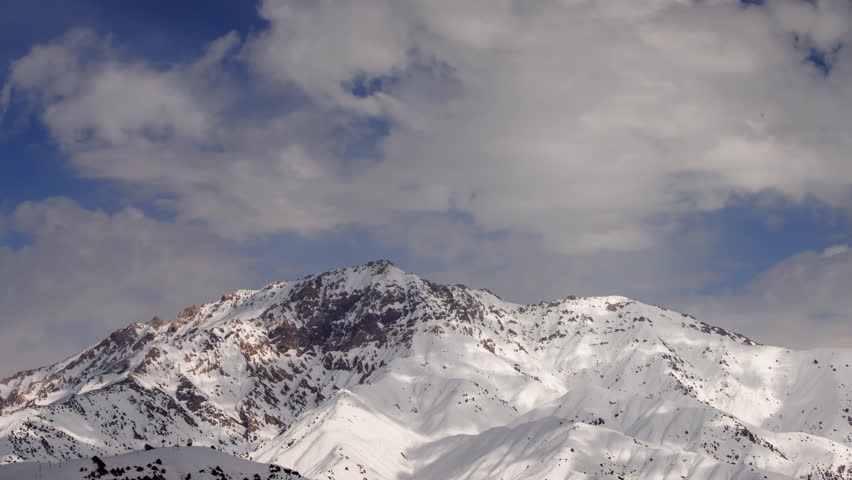 Winter mountains in snowy timelapse. Blue clouds fly over the snow-covered peak. Sun brightly shines in white snow. Cinematic video for background. Atmosphere of amazing nature, breathtaking