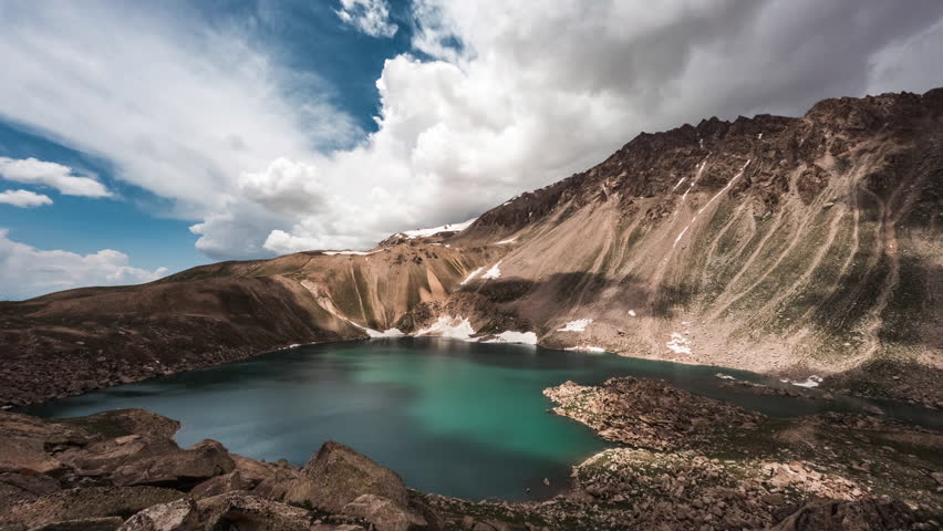 Mountains and lake video timelapse for background. A small lake on top of Mount Arashan. Over the lake in blue sky run heavy cumulus clouds. Remains of snow at water's edge. Joyful peaceful cinematic