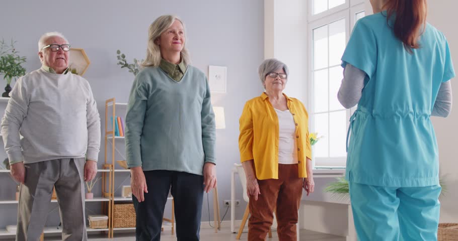 Young nurse or caregiver doing healthy exercises for wellness and health with a group of seniors. Elderly people working out in fitness class of nursing home with female physiotherapist. 4k video.