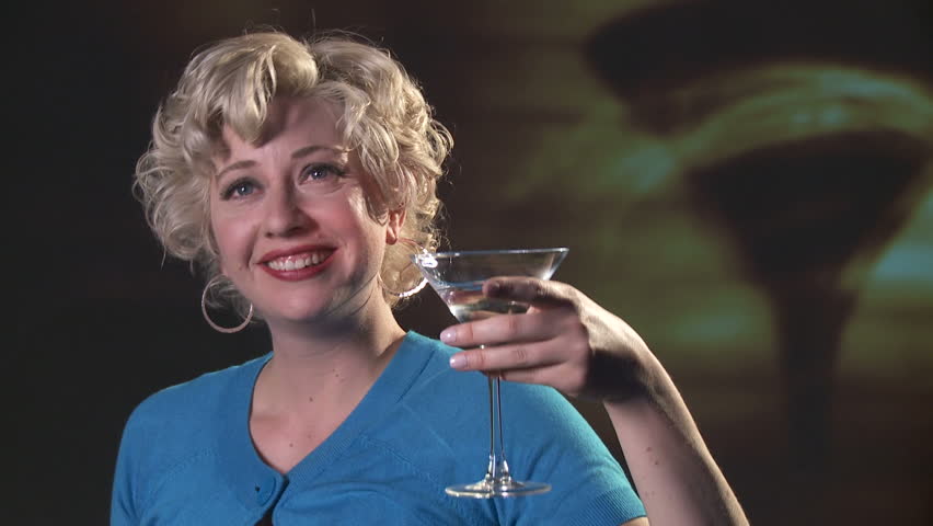 Blonde girl in a turquoise dress holds a martini in front of a large shadow of a