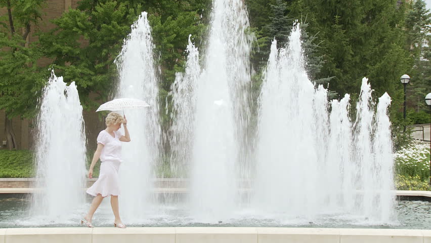 Blonde girl with a polka dot umbrella walks along the edge of a fountain, stops
