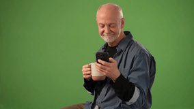 Mature man smiles as he sips from a mug while viewing his smartphone, surrounded by a solid green background, ideal for announcements or product presentation. - Powered by Shutterstock - Get 15% off with code: PIKWIZARD15