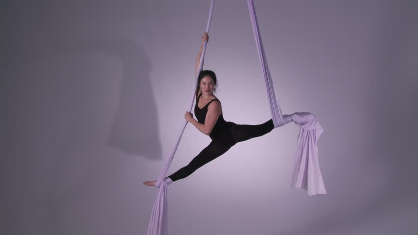 Gymnastic young woman does splits and spins while doing aerial yoga. Wide shot