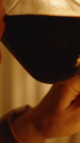 Vertical video. Woman Sipping Red Wine from Glass in Warm Ambient Light with Focus on Fingers and Stem of Glass, Evening Relaxation Concept