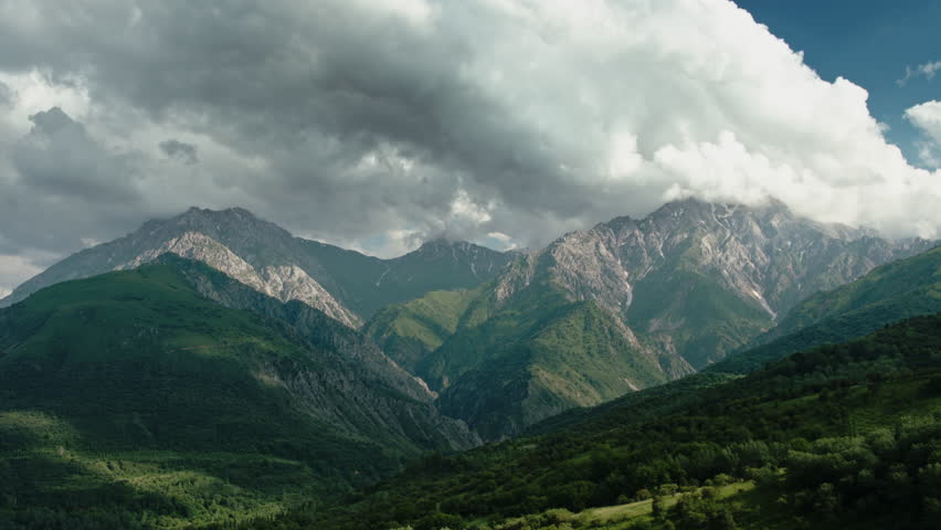 Mountain landscape timelapse video for background. Rolling mountains covered with green vegetation and rocky mountains with remnants of snow in background under blue sky with swirling clouds. Alpinism