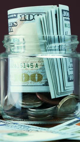 Rolled bills of US dollar banknotes and coins in glass jar rotating on violet background. Rolled one hundred dollars are in moneybox. Money saving concept. Vertical video