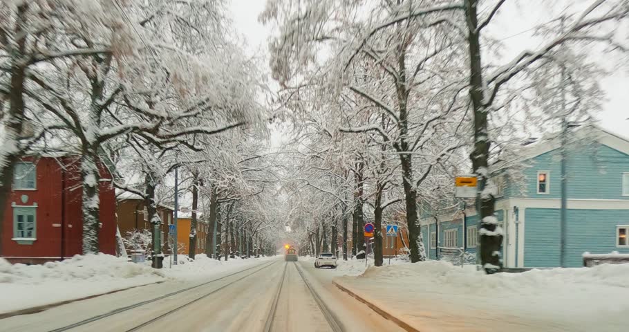 POV view from car driving on Pohjolankatu street and passing tram track cleaning trucks in cloudy winter weather with snow on the ground, Helsinki, Finland.