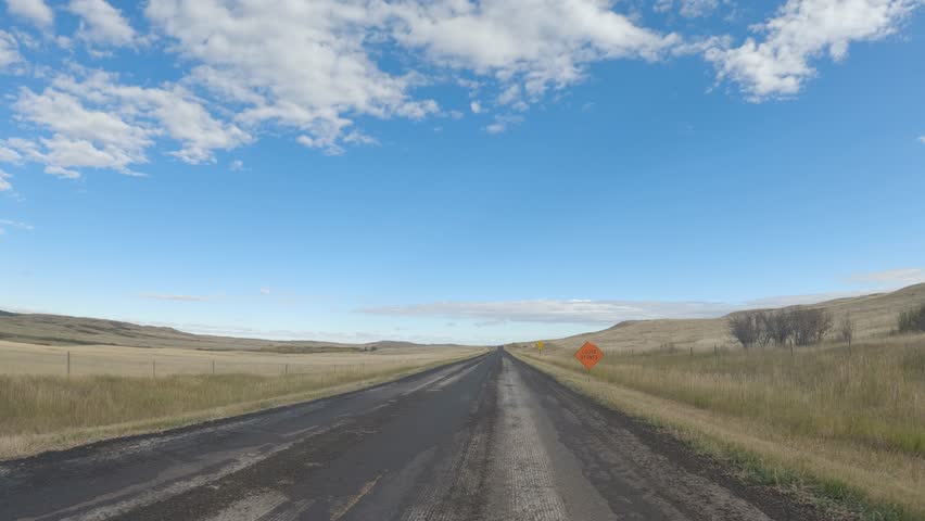 Point of view driving along a rough paved road through rolling ranch land under a blue sky with scattered clouds