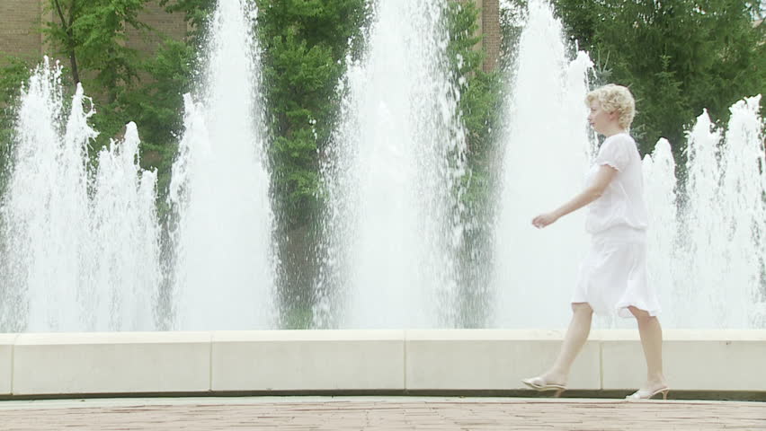 Blonde girl walks past a fountain, turns and waves at someone unseen, then