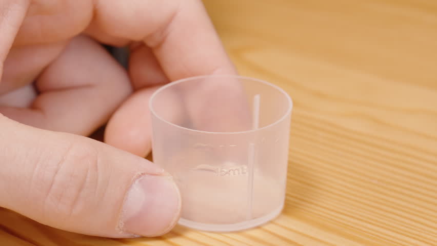 Close-Up of Liquid Being Poured from Brown Bottle into Measuring Cup with Green Solution on Wooden Table Liquid Medicine Usage