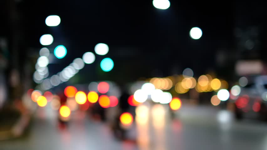 A blurry image of a busy street at night with cars and motorcycles. The lights from the vehicles and street lamps create a sense of movement and energy