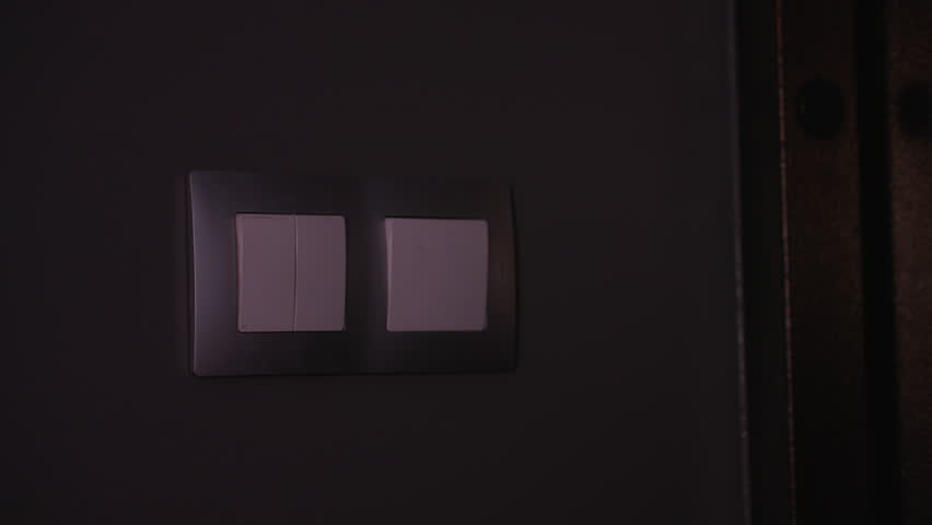Close-up of woman hand pressing light switch in dimly lit room, soft ambient glow highlights fingers as she turns on the light, creating a contrast between darkness and illumination