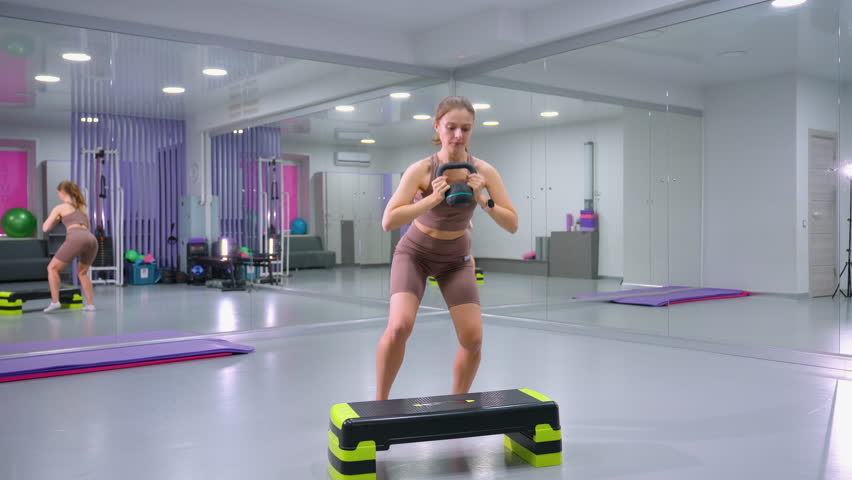 Woman lifts weight, steps onto aerobic bench, then steps down and lifts weight again in a gym with mirrored walls, strength training and cardio exercise routine combining muscle-building