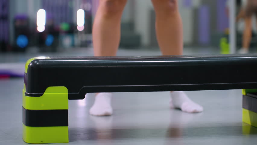 Close-up lower angle view of individual stepping onto exercise bench and stepping down during workout, gym environment with fitness equipment in background