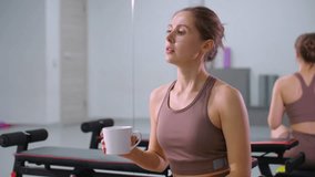 Woman takes break from fitness session, sipping coffee while seated in gym, large mirror reflects relaxed posture, workout equipment and mats in background - Powered by Shutterstock - Get 15% off with code: PIKWIZARD15