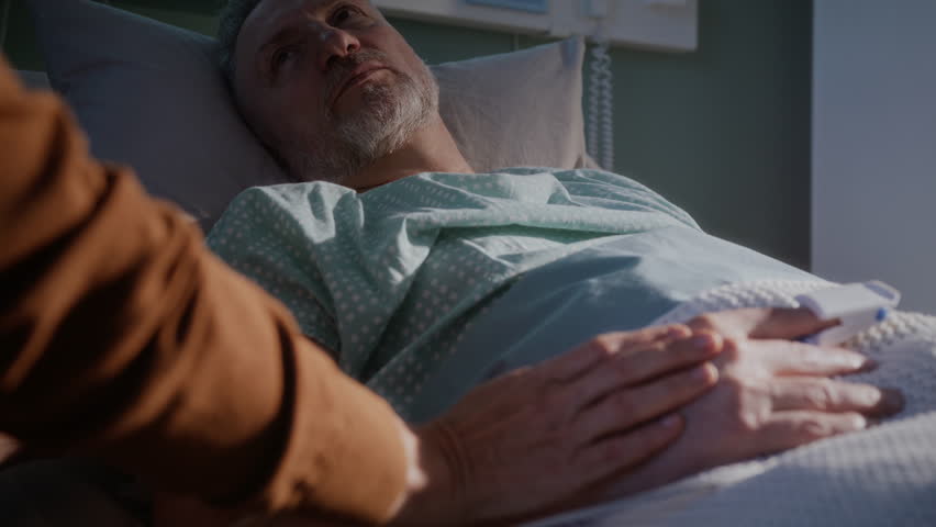 Senior man with grey beard looks at supportive wife holding hands in hospital bed. Mature couple shares love while weak husband rests after surgery