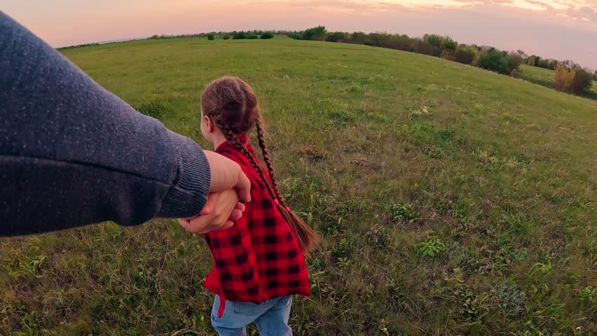 Happy kid girl holds father's hand runs on grass. Girl child holds daddy's hand. Dad child walk on grass in park holding hands. Happy family. Father with little daughter together on meadow at sunset.