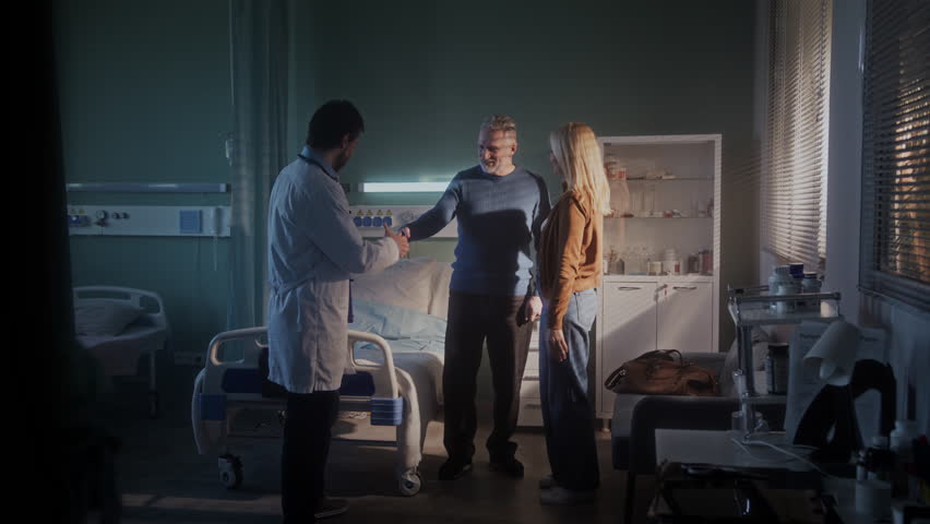 Male doctor in white coat interacts with senior man with gray hair standing near woman. Patient hugs family member looking grateful ready for discharge