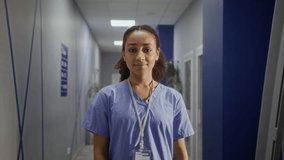 Female medical professional smiles broadly standing in hospital corridor. Young woman doctor wearing blue scrubs indicating role in medical field - Powered by Shutterstock - Get 15% off with code: PIKWIZARD15