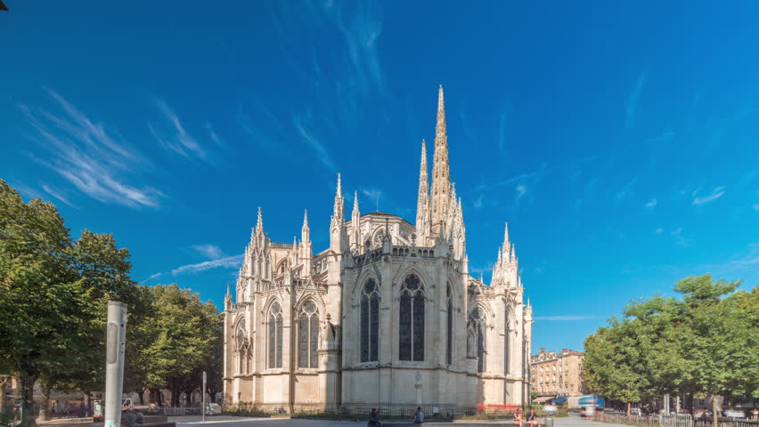 Cathedrale Saint-Andre de Bordeaux timelapse hyperlapse with twin spires under a vibrant blue sky. Green trees and people walking in the square create a serene urban atmosphere. Bordeaux, France