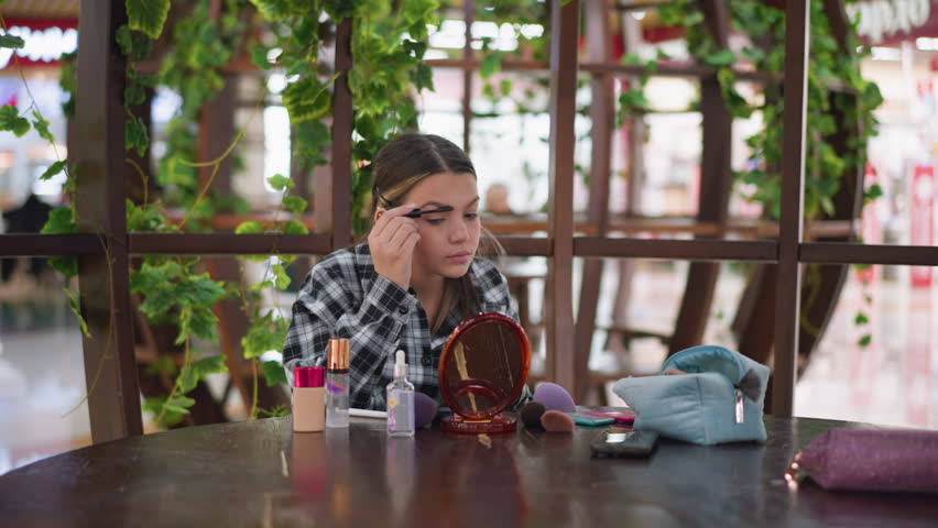 Close-up shot of young woman applying makeup on her eyebrows with eyebrow pencil and brush, using a mirror, various makeup products such as foundation and brushes placed on table