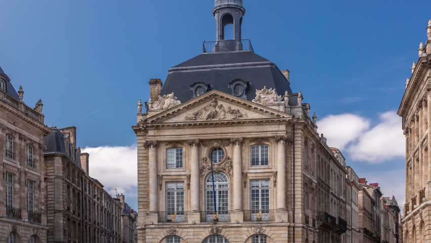 Place de la Bourse timelapse hyperlapse in Bordeaux, France, with restaurants surrounded by classic architecture under a cloudy blue sky. People walking around the main square and central streets