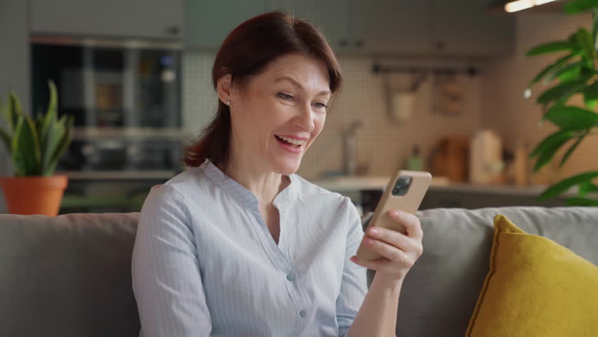excited middle aged woman saying wow while looking at smartphone screen. happy moment of surprise and joy on cozy couch in stylish modern kitchen, natural light, real-life reactions at home