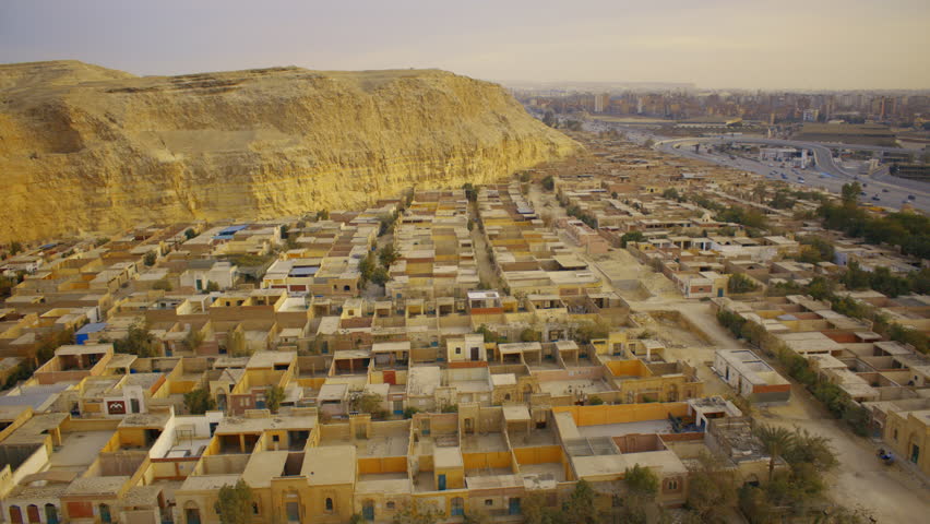 An aerial view of an old cemetery in Cairo, featuring tightly packed tomb structures in sand-colored hues, narrow pathways, and sparse vegetation. City skyline and highway visible in the background.
