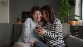 smiling mom and teenage daughter with acne bonding over funny content on smartphone. cozy moment on sofa bright kitchen, relaxed weekend vibes, real family connection at home. video call with family - Powered by Shutterstock - Get 15% off with code: PIKWIZARD15
