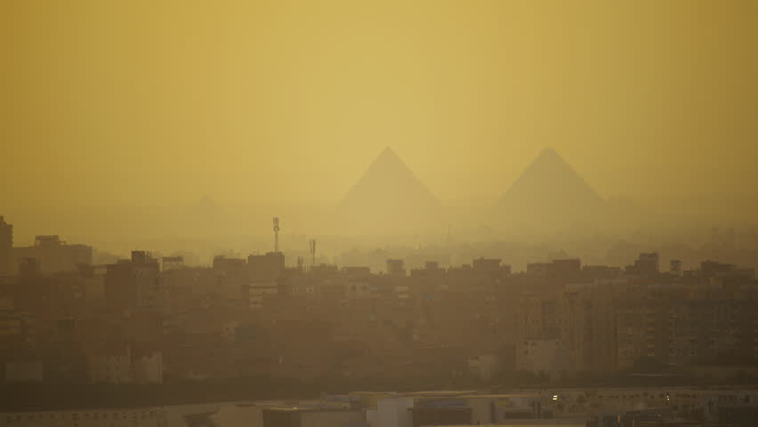 Overview of Cairo skyline with pyramids of Giza silhouetted against a glowing yellow sky in the mist and dusty air. Majestic Egyptian pyramids tower above the Cairo cityscape.