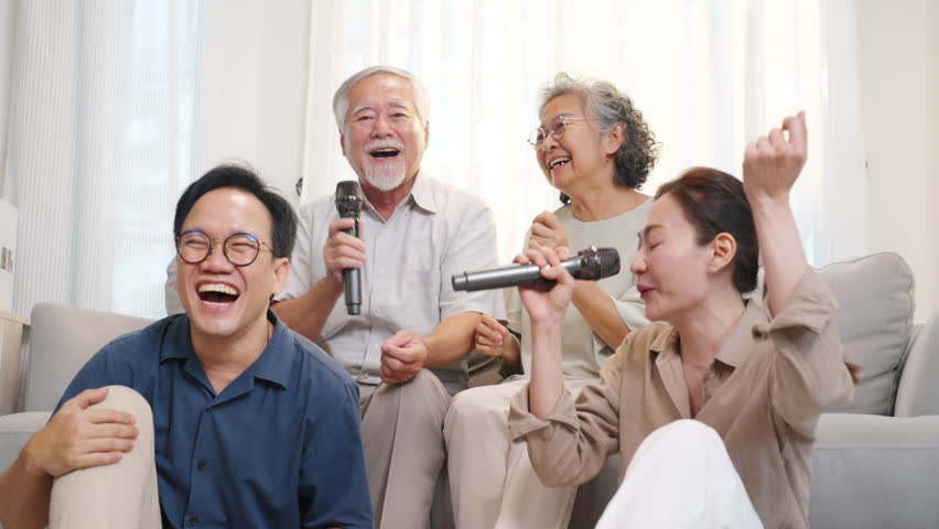 Happy Asian family senior parents and adult child spending time together at home. Healthy elderly father and mother enjoy and fun singing karaoke song dancing with daughter and son in living room.