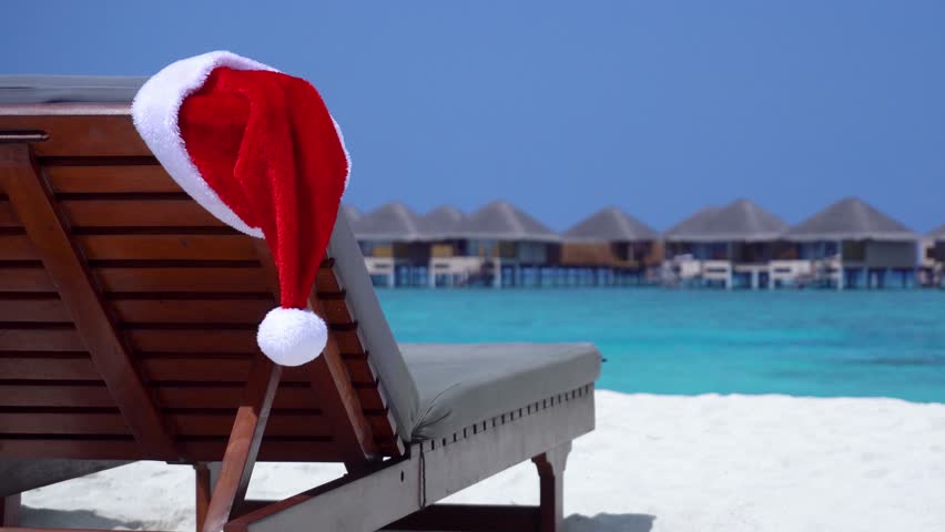 Santa Claus Helper Hat on chair at sandy beach with turquoise sea and water bungalows on background. Christmas celebration in tropical destinations