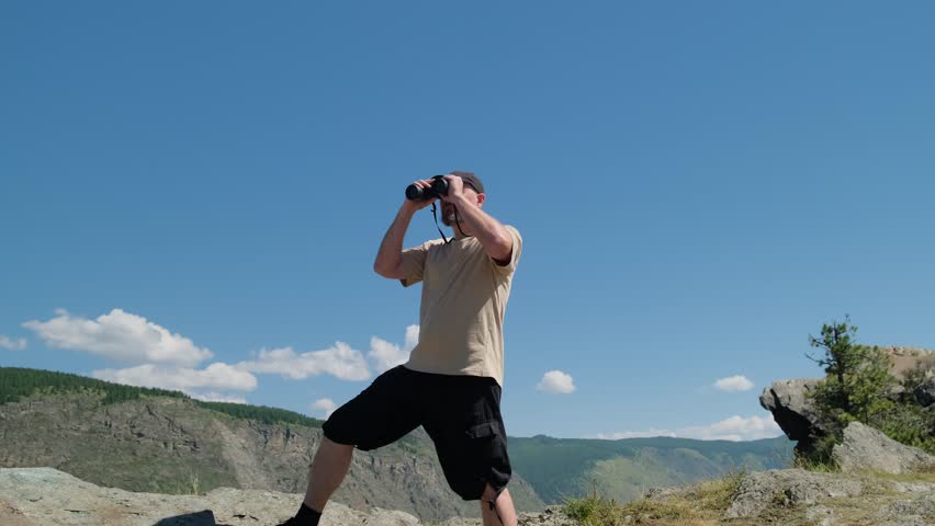 A man traveler looking through binoculars standing on top of mountain with blue sky on the background.
