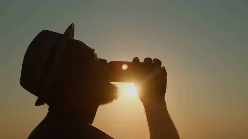 Adult bearded man in a hat drinking beer from a can by the sea at sunset. A refreshing sip of cold drink on a hot summer evening on vacation.