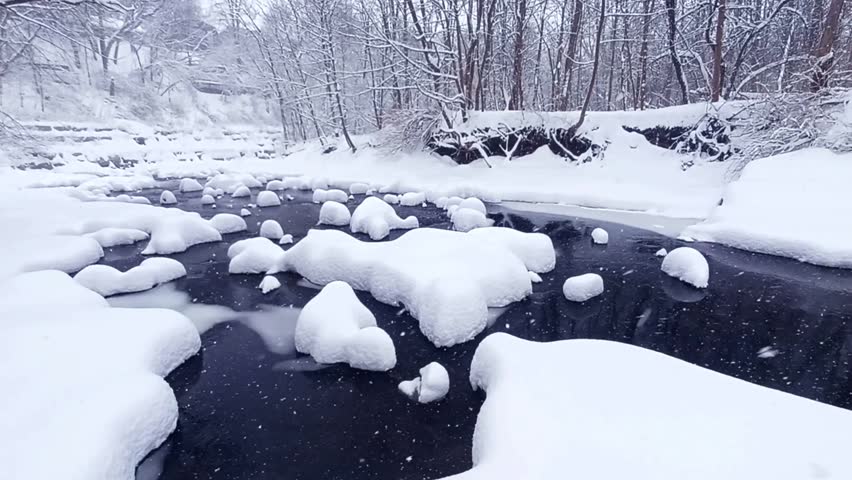 A  serene creek flows through a forest, with rocks and banks blanketed in deep snow during a heavy snowfall.