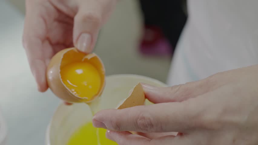 Close-up shot of hands skillfully separating yolk from egg white in a kitchen setting, illustrating cooking preparation. Close-Up of Hands Separating Egg Yolk in Kitchen