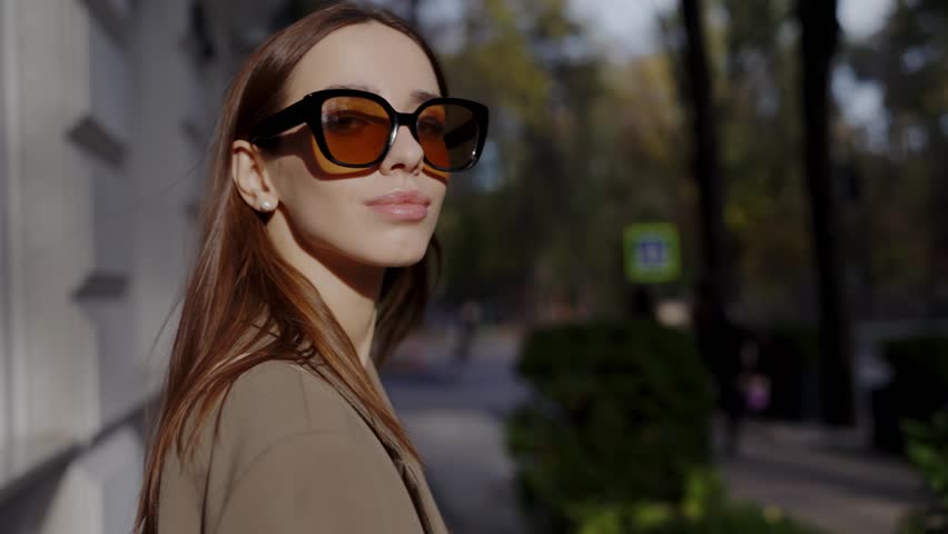 Chic young woman wearing stylish sunglasses enjoys a sunny day on a bustling city sidewalk. A perfect blend of fashion and urban lifestyle. Elegant Woman in Sunglasses Walking in a Sunlit City Street