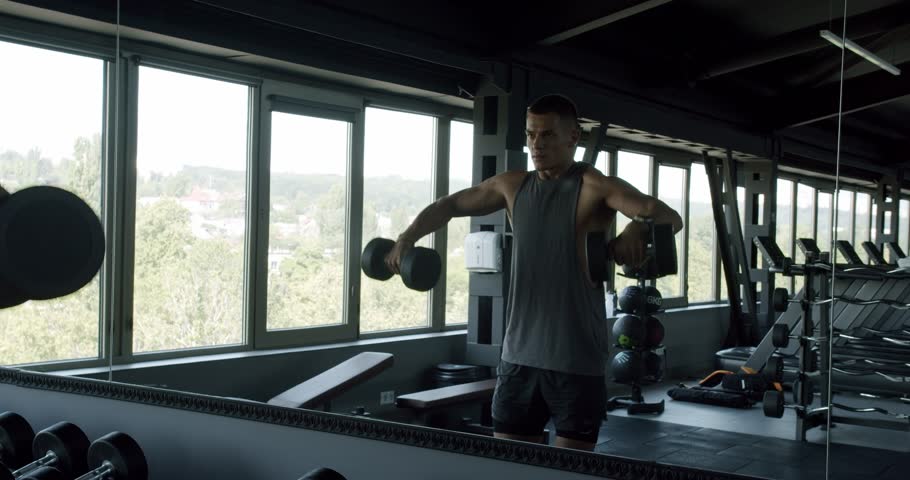 A muscular man in a tank top lifting dumbbells in a bright, modern gym with large windows showing a sunny landscape. Focused Man Lifting Weights in a Modern Sunny Gym