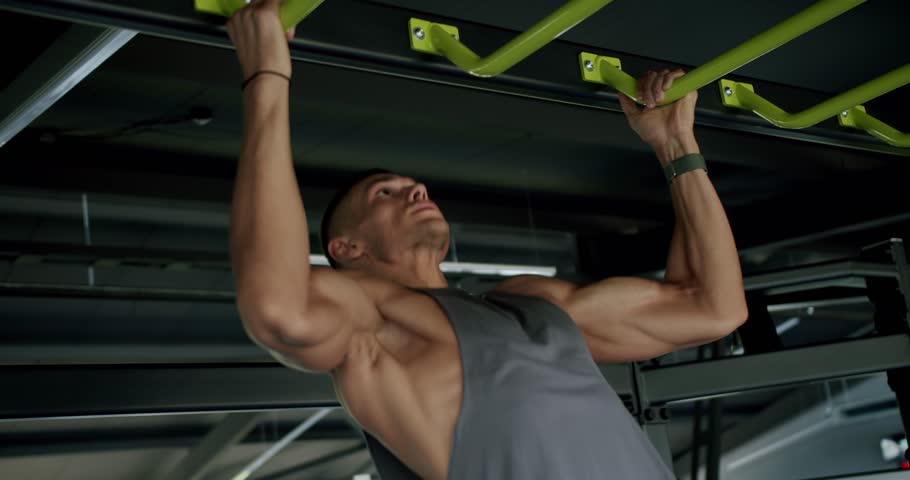 Focused Man Performing Challenging Pull-Ups in Modern Fitness Gym. Fit male athlete engaging in intense pull-up exercise on a green bar in a well-equipped, contemporary gym setting.
