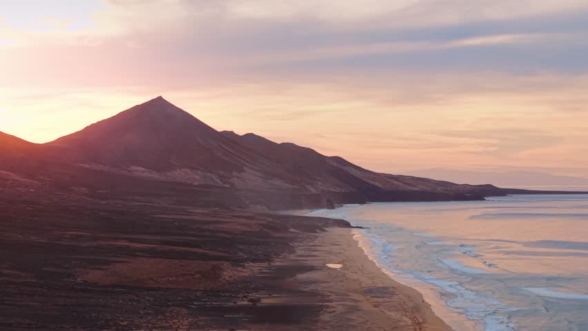 Slow circular drone pan over Cofete beach in Fuerteventura during golden hour, with warm sunlight, deep blue sea, gentle waves, and the sun setting behind mountain peaks