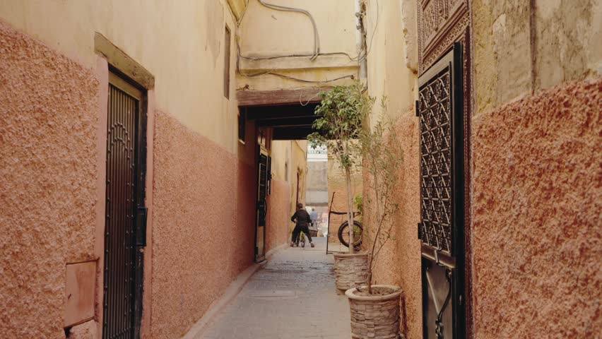 A young boy rides his bike down a narrow alley in Marrakech, Morocco. The walls are painted a warm, inviting shade of light orange. Plants in pots add a touch of greenery to the scene.