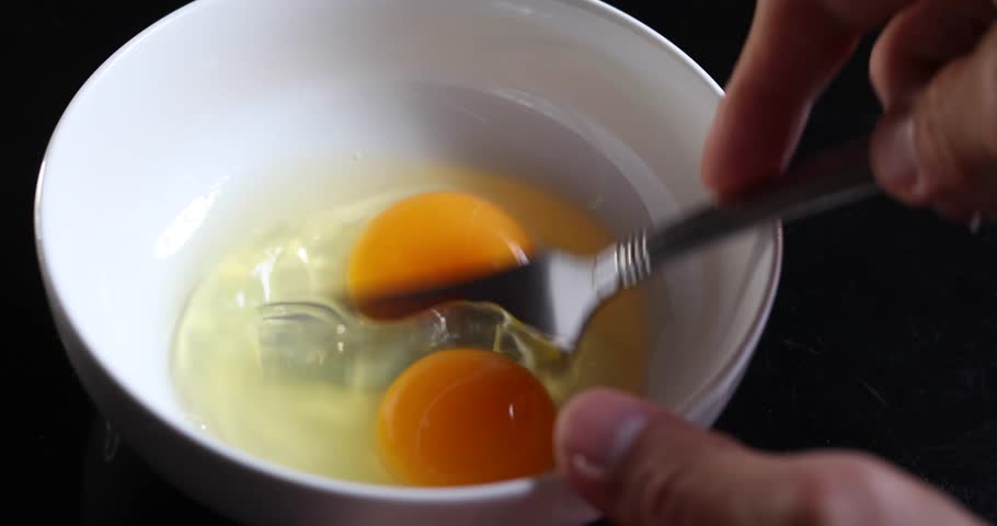 Close-up of raw eggs being beaten in a white bowl with a fork. The yolk and whites are still separated while the fork mixes them together. 