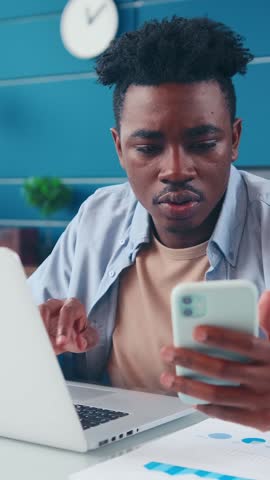 Young African American man sits at a clean workspace, focused on his smartphone. He gestures with his hand as he is engaged in a lively conversation, possibly sharing ideas or solutions.