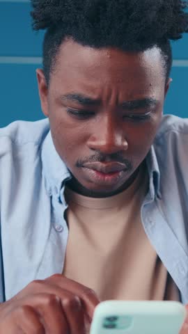 In a vibrant setting, young African American man intensely browses his phone, showcasing focus and curiosity. His expression reflects a mix of determination and thoughtfulness.