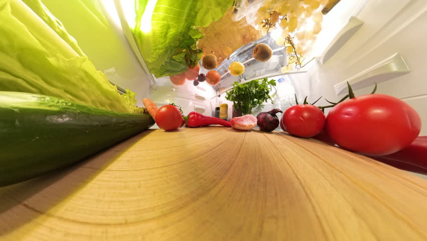 The refrigerator door opens as a man takes and eats a piece of peeled grapefruit. The foreground shows fresh produce like tomatoes, onions, and peppers, adding vibrancy to the scene
