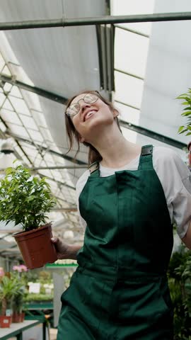 A cheerful brunette girl in glasses and a green apron dances with houseplants in her hands in a greenhouse